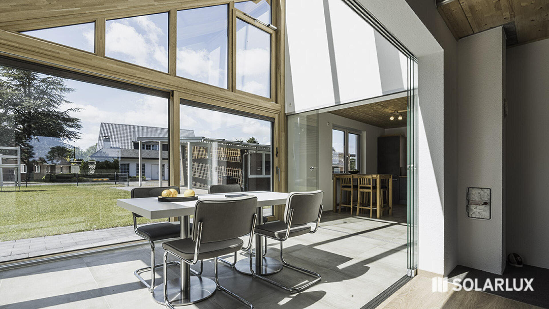 Moderner Essbereich mit großen Glasfenstern und Glasschiebetüren, die den Blick auf einen grasbewachsenen Hof und die benachbarten Häuser freigeben. Das Sonnenlicht erfüllt den Raum. Das SOLARLUX-Logo ist in der unteren rechten Ecke zu sehen.