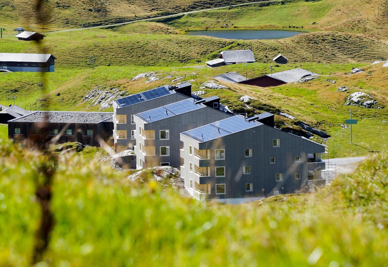 Drei moderne Wohngebäude mit Sonnenkollektoren auf den Dächern liegen auf einem grasbewachsenen Hügel, umgeben von verstreuten kleinen Strukturen und einer sanften grünen Landschaft im hellen Sonnenlicht.