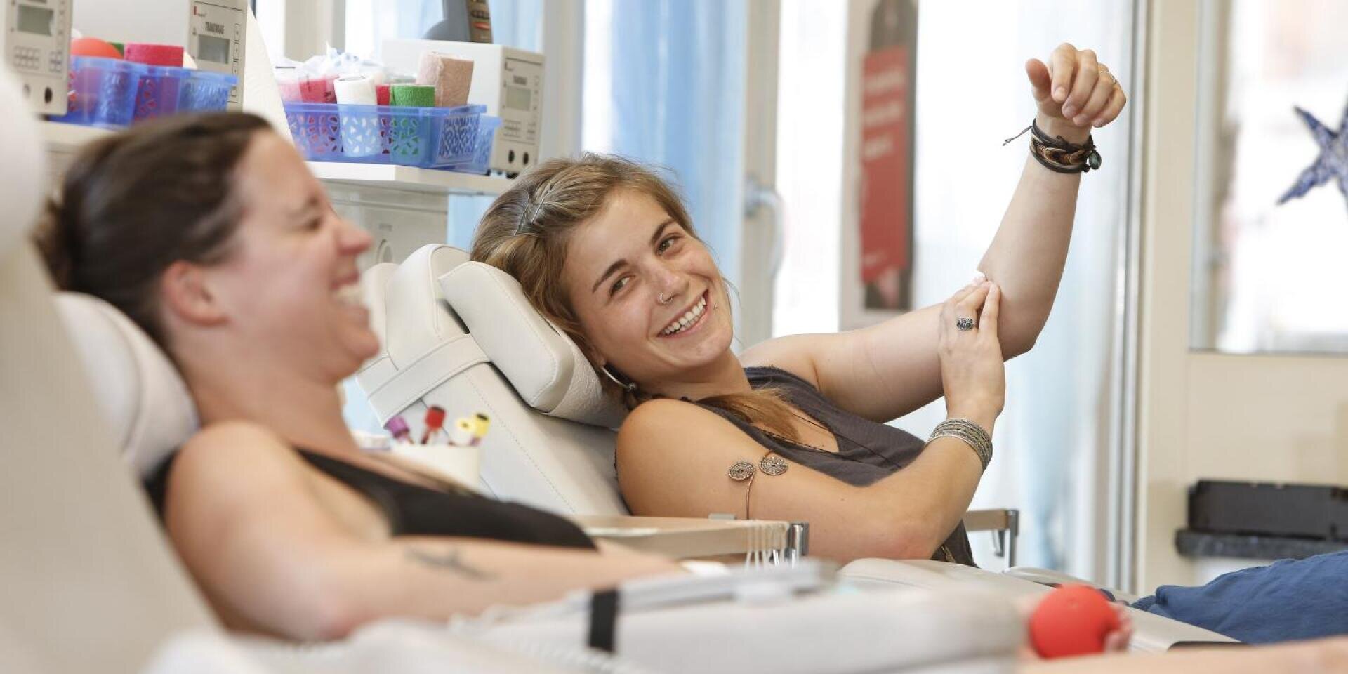 Zwei Frauen sitzen auf Stühlen, lächeln und lachen, während sie in einer hellen, modernen Klinik Blut spenden. Eine Frau zeigt auf ihren Arm, in den eine Nadel eingeführt wird, und im Hintergrund sind Spenderutensilien zu sehen.