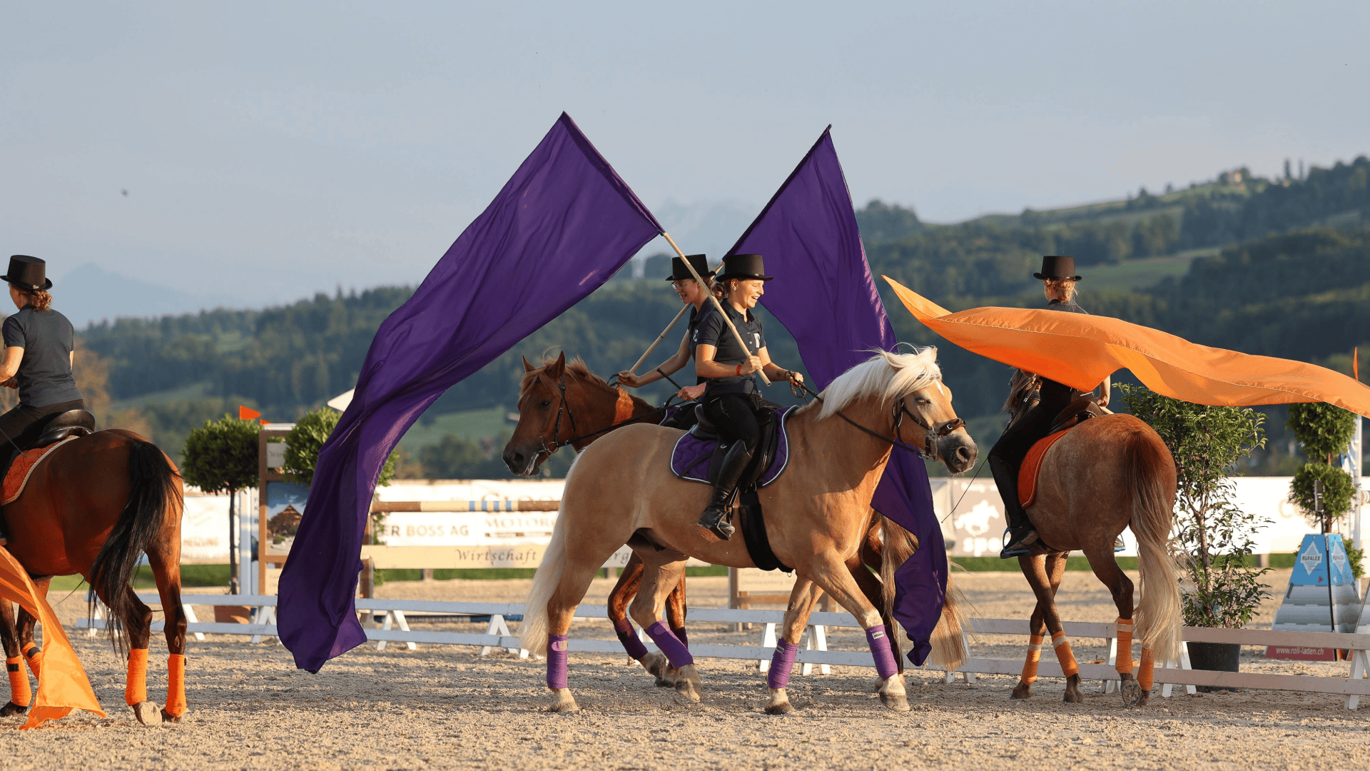 Drei schwarz gekleidete Personen mit Zylinder reiten auf Pferden in einer Freiluftarena. Zwei von ihnen tragen große, wehende violette Fahnen, eine hat eine orangefarbene Fahne. Die Pferde haben passende Beinkleider. Im Hintergrund sind Hügel und Bäume zu sehen.