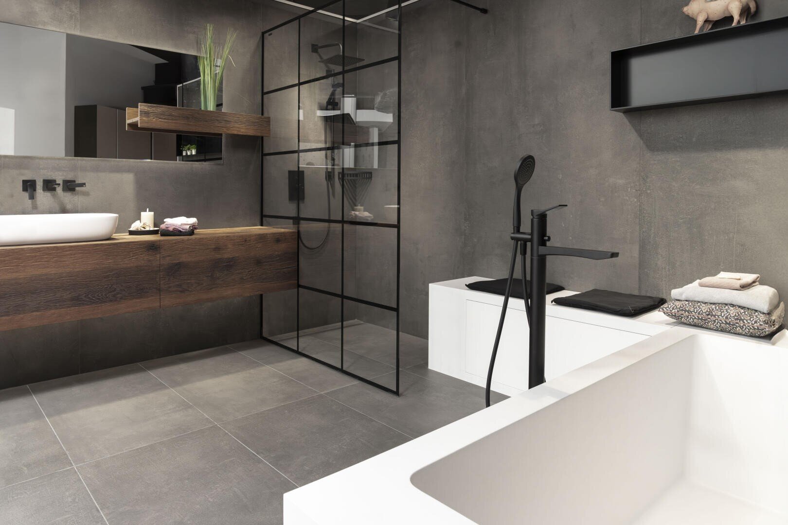 Modernes Badezimmer mit grauen Wänden und Böden, einer freistehenden weißen Badewanne, einem schwebenden hölzernen Waschtisch mit Waschbecken, einer Glasduschabtrennung, schwarzen Armaturen und ordentlich gestapelten Handtüchern. Minimalistisches und stilvolles Design.