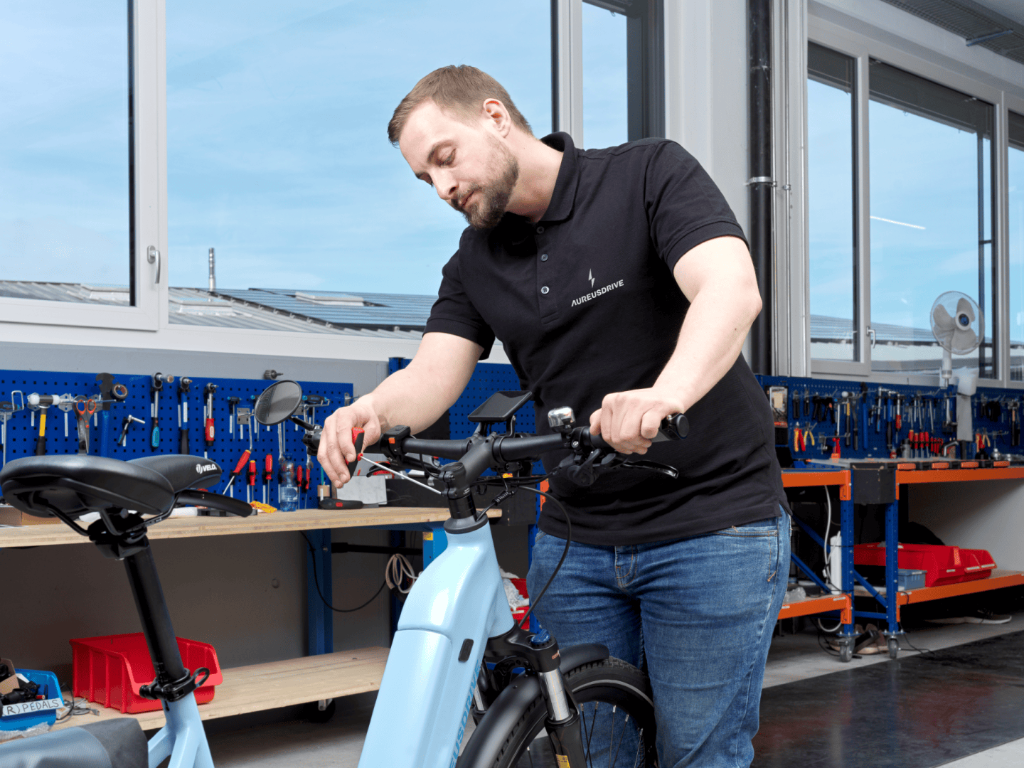 Ein Mann in einem schwarzen Polohemd stellt den Lenker eines hellblauen Elektrofahrrads in einer Werkstatt ein, in der im Hintergrund Werkzeuge und Geräte in Regalen stehen.