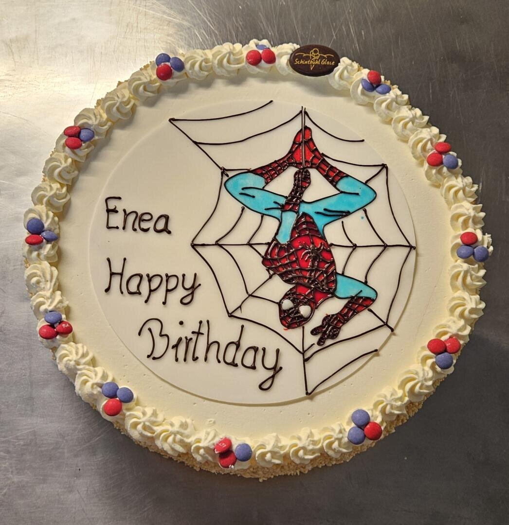 Ein runder Geburtstagskuchen mit weißem Zuckerguss, verziert mit einer Zeichnung von Spider-Man, der kopfüber in einem Netz hängt, und der Botschaft Enea Happy Birthday. Der Kuchen hat Sahne und bunte Süßigkeiten Dekorationen rund um den Rand.