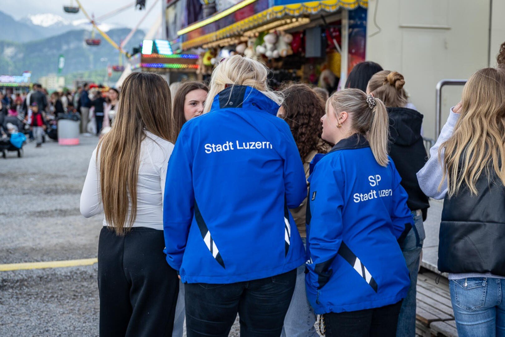 Eine Gruppe von Frauen steht auf einem Jahrmarkt zusammen. Zwei tragen blaue Jacken mit der Aufschrift Stadt Luzern. Im Hintergrund sind bunte Fahrgeschäfte und Berge zu sehen. Die Frauen sind von hinten zu sehen, in ein Gespräch vertieft.