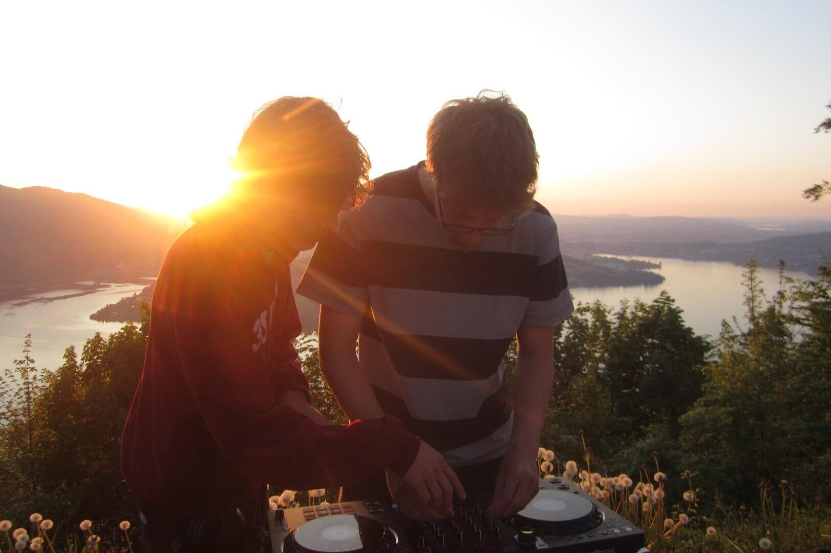 Zwei Personen stehen bei Sonnenuntergang im Freien an einem DJ-Mischpult. Zwischen ihnen scheint das Sonnenlicht, und im Hintergrund ist ein malerischer Blick auf einen Fluss, Hügel und Bäume zu sehen.