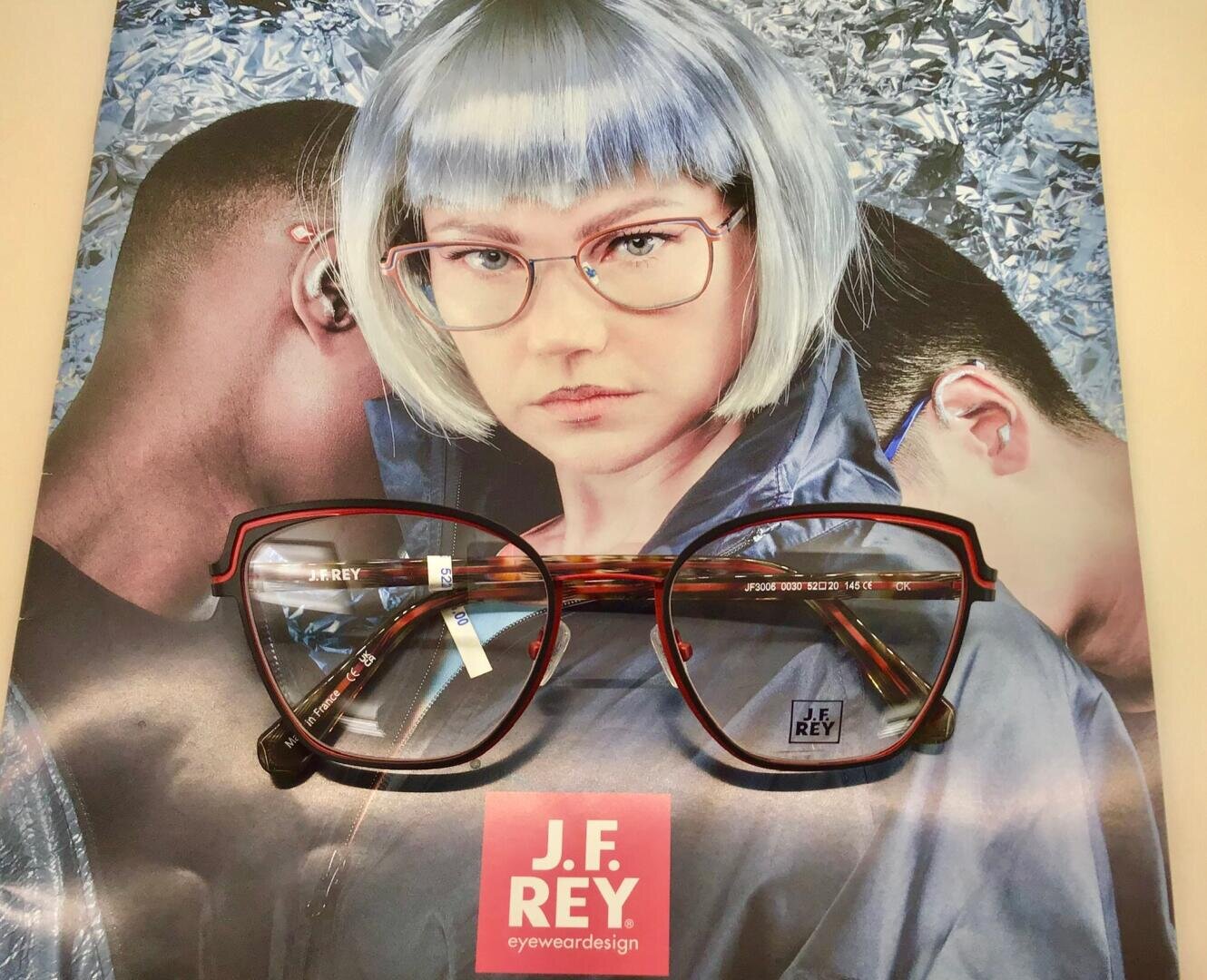 Nahaufnahme einer J.F. Rey-Brille auf einem Plakat, das eine Frau mit kurzem silbernem Haar und rotgerahmter Brille zeigt, flankiert von zwei Personen, die sich abwenden, mit glänzender Silberfolie im Hintergrund.