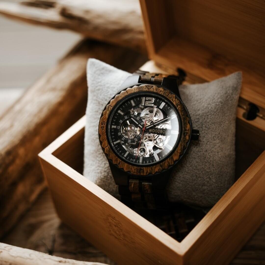 Eine hölzerne Armbanduhr mit einem sichtbaren mechanischen Uhrwerk liegt auf einem grauen Kissen in einem offenen Holzkasten auf einer Holzfläche.