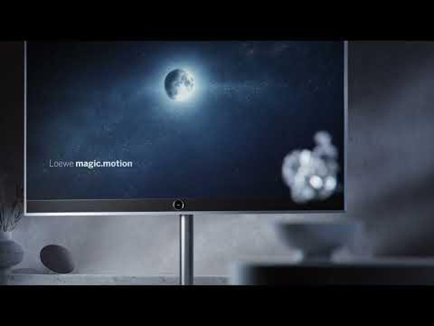 Ein moderner Flachbildfernseher zeigt ein Bild des Mondes in einem dunklen Himmel mit der Aufschrift Loewe magic.motion; der Raum ist schwach beleuchtet, im Vordergrund sind unscharfe Objekte zu sehen.
