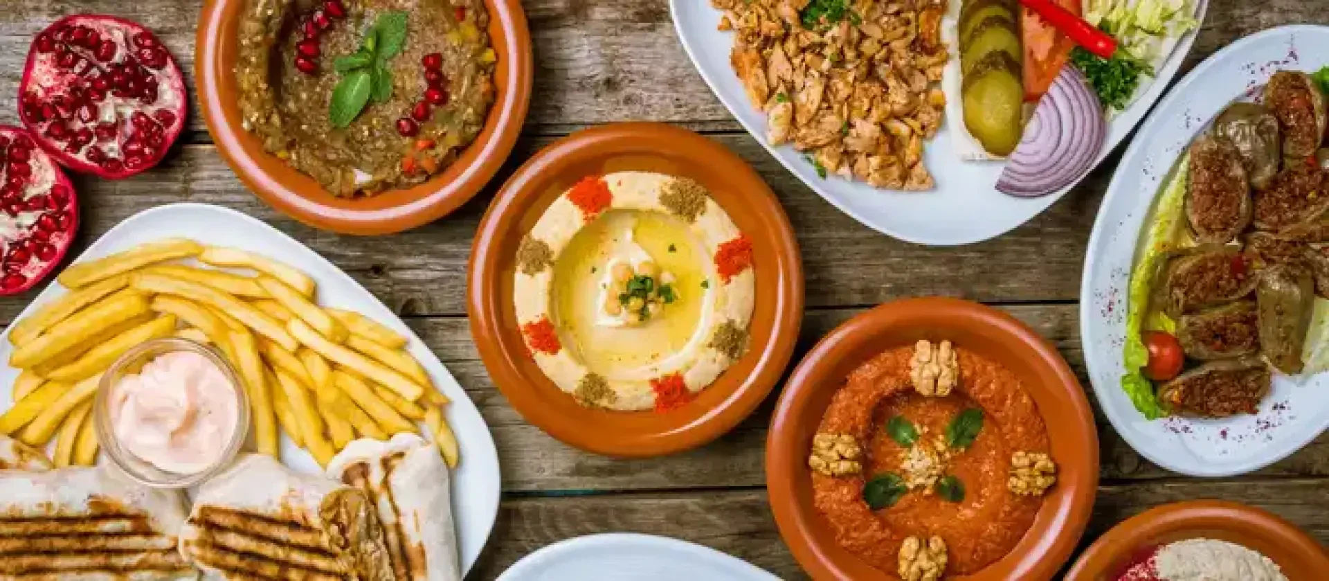 Auf einem rustikalen Holztisch stehen bunte orientalische Gerichte wie Hummus, Baba Ghanoush, Pommes frites, gegrilltes Fleisch, Salate, gefülltes Gemüse und frische Granatapfelhälften.