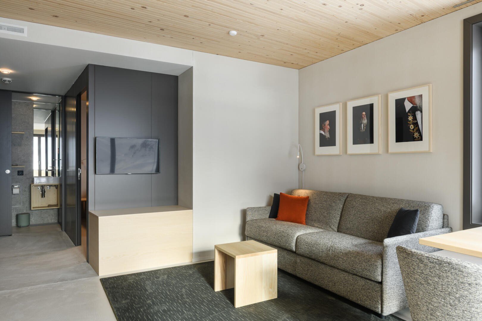 Ein modernes Hotelzimmer mit einem grauen Sofa, einem orangefarbenen Kissen, einem Couchtisch aus Holz, einem an der Wand montierten Fernseher, gerahmten Porträts an der Wand und einer hellen Holzdecke. Große Fenster lassen viel natürliches Licht herein.