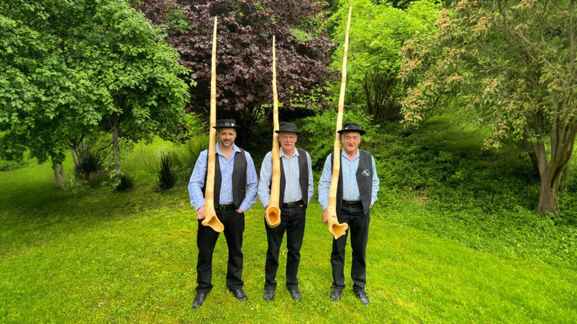 Drei Männer mit Hüten und Westen stehen im Gras und halten lange hölzerne Alphörner senkrecht, umgeben von üppigen grünen Bäumen und Büschen.