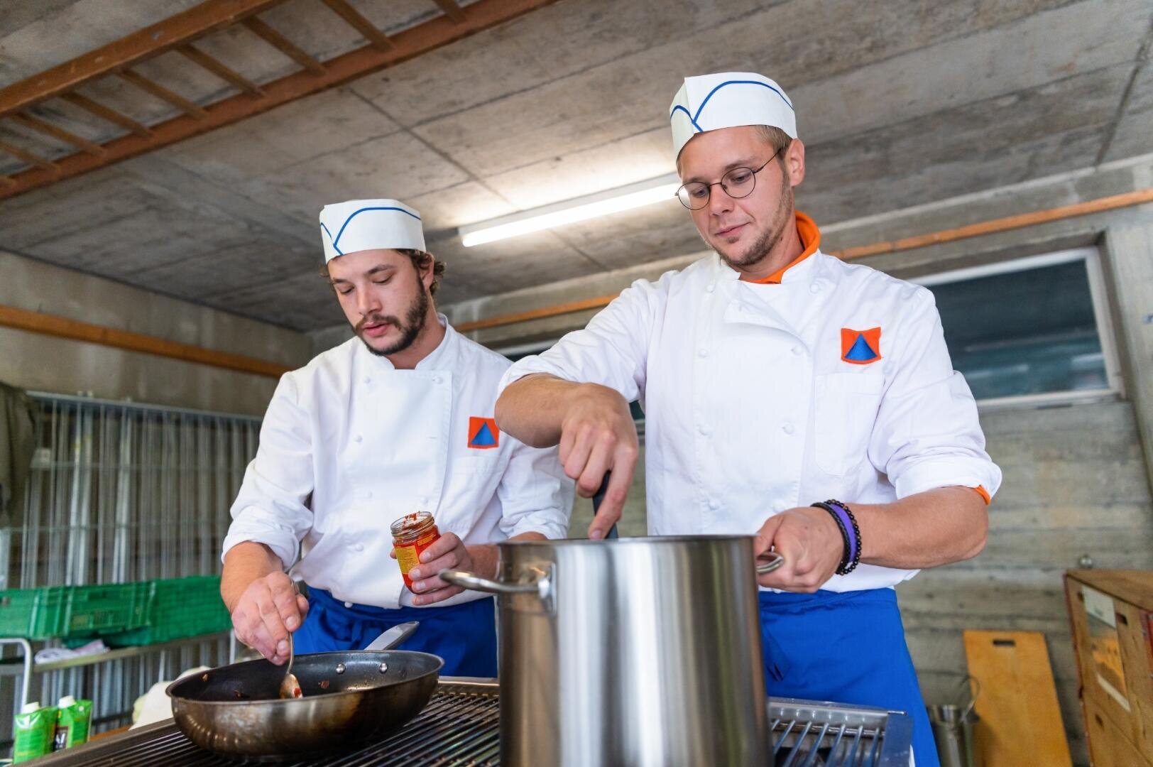 Zwei Köche in weißen Uniformen und Mützen kochen gemeinsam; einer rührt in einem großen Topf, während der andere in einer Großküche Zutaten in eine Pfanne gibt.