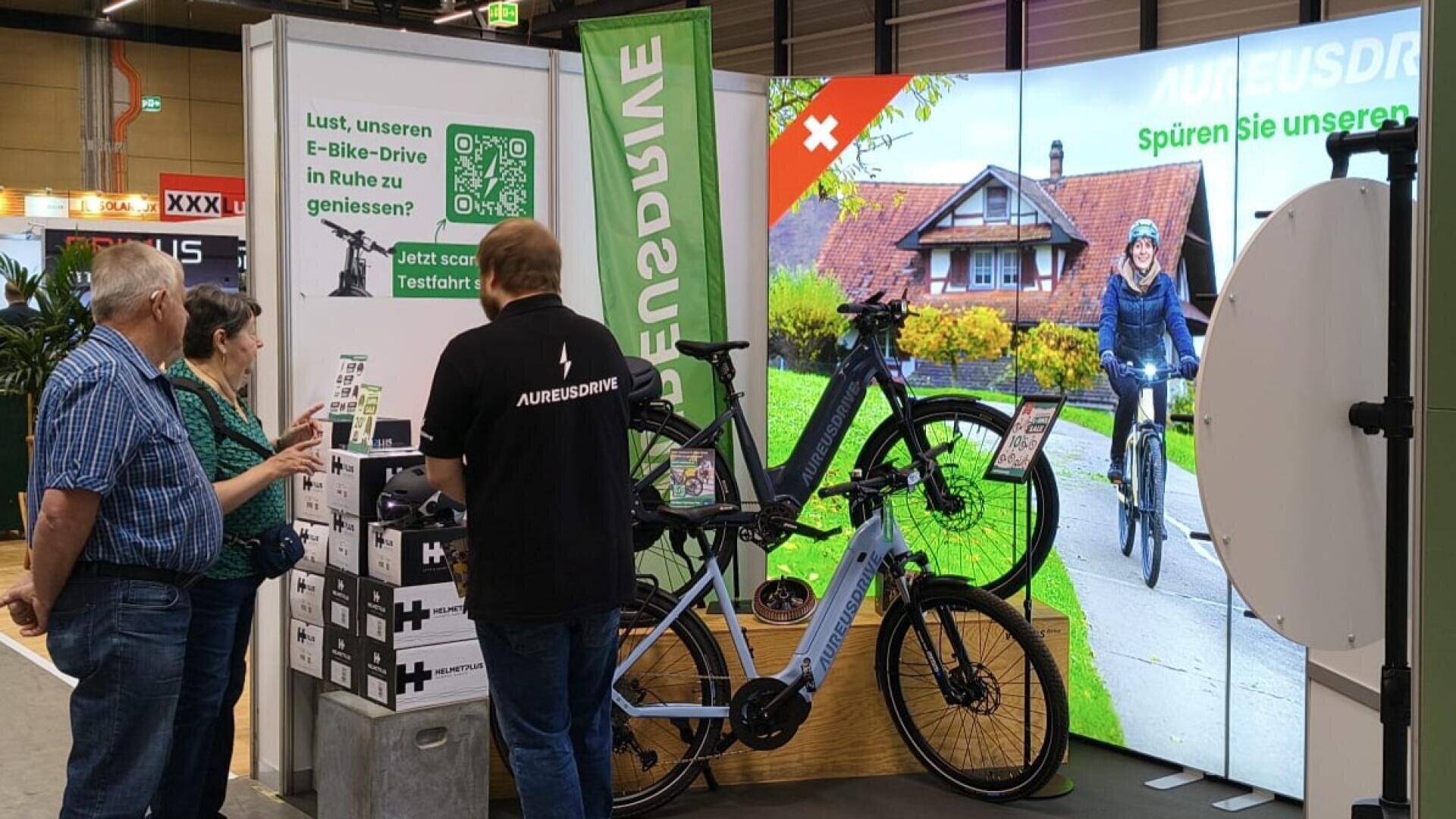 Ein Mann und eine Frau unterhalten sich mit einem Verkäufer an einem AurusDrive-Stand mit ausgestellten E-Bikes. Ein großer Bildschirm zeigt einen Radfahrer, der an einem Haus vorbeifährt und für die Marke wirbt. Ein QR-Code und grüne Banner sind zu sehen.