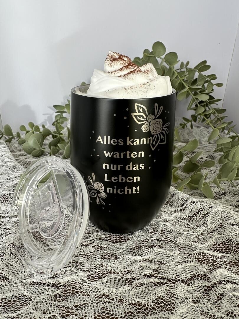 Ein schwarzer Becher mit floralen Motiven, der mit Schlagsahne und Kakaopulver gefüllt ist, steht auf einem Spitzenstoff, daneben ein durchsichtiger Deckel. Der deutsche Text auf dem Becher lautet: "Alles kann warten nur das Leben nicht! Grünzeug ziert den Hintergrund.