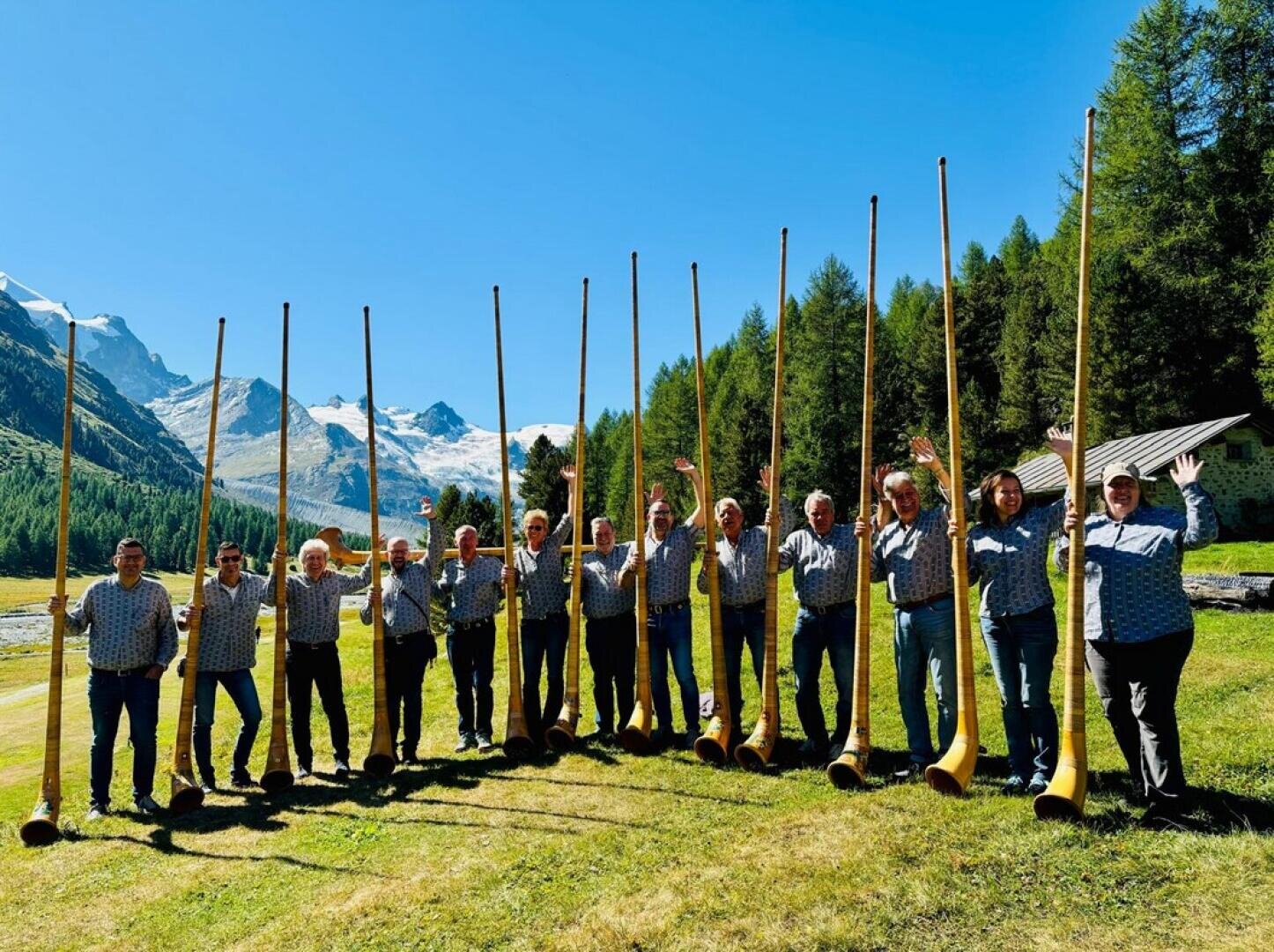 Zwölf Personen stehen im Freien auf einer Wiese und halten jeweils ein langes Alphorn aufrecht. Im Hintergrund sind schneebedeckte Berge, Bäume und der blaue Himmel an einem sonnigen Tag zu sehen.