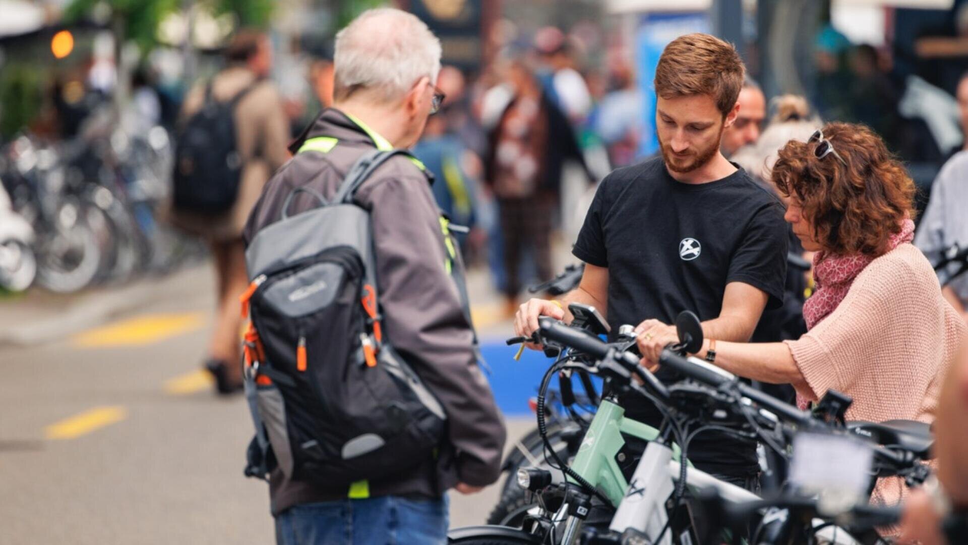 Ein junger Mann hilft zwei Erwachsenen bei der Untersuchung von Fahrrädern bei einer Veranstaltung im Freien. Die Erwachsenen sehen sich die Fahrräder genau an, während andere Menschen und weitere Fahrräder im geschäftigen Hintergrund verschwommen erscheinen.