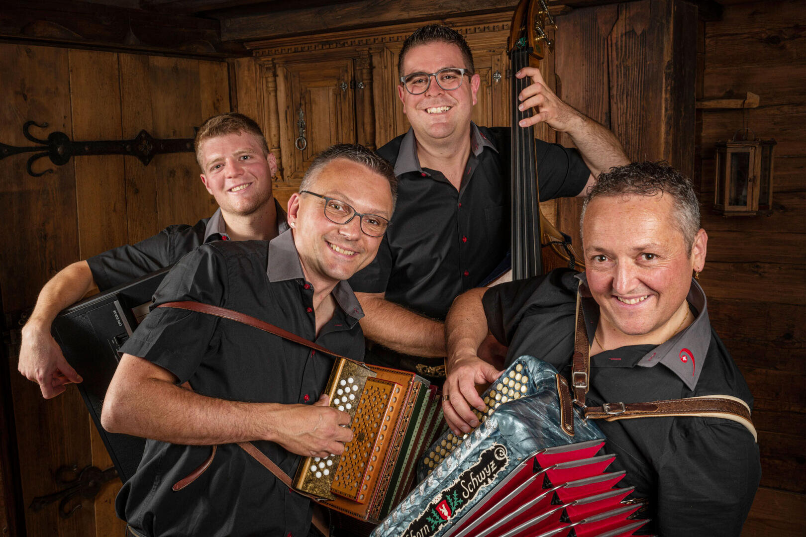Vier lächelnde Männer in passenden schwarzen Hemden posieren mit Musikinstrumenten, darunter ein Akkordeon, ein Kontrabass und eine Konzertina, in einem rustikalen Holzzimmer.