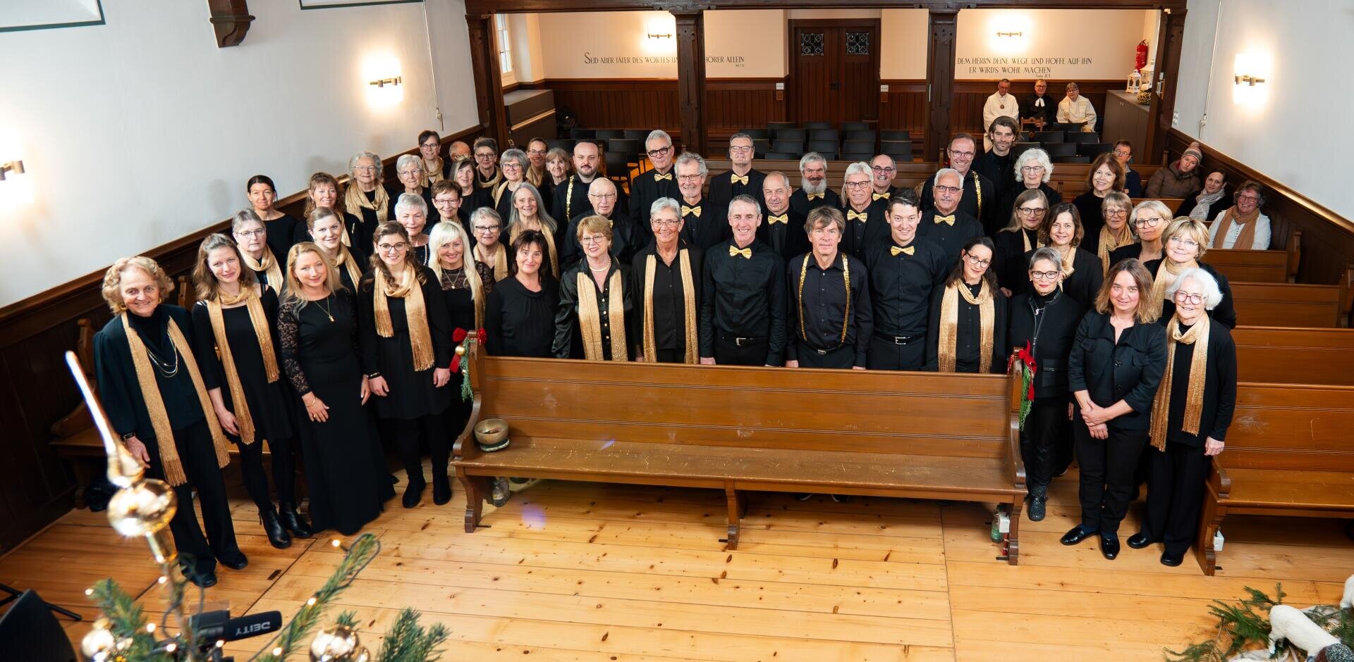 Ein großer, schwarz gekleideter Chor mit goldenen Schals und Fliegen steht in einer Holzkirche und lächelt in die Kamera. Einige halten Rosen. Vorne sind die Kirchenbänke leer und hinten sitzen ein paar Menschen.