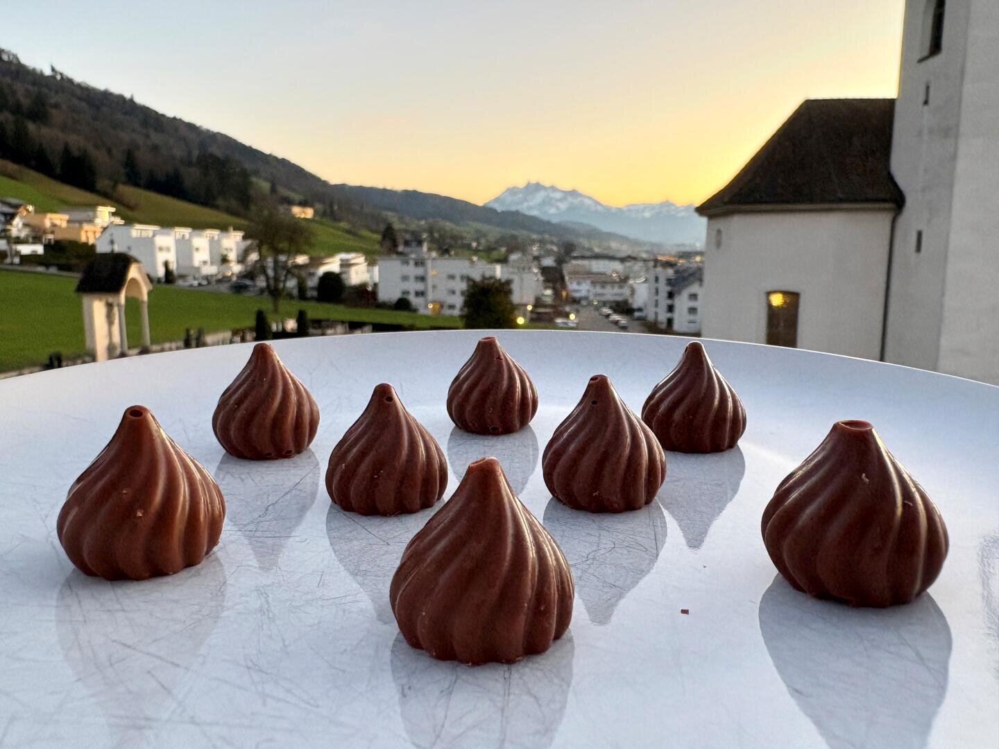 Acht Bonbons in Form von Schokoladenbaisern liegen auf einem weißen Teller im Freien, vor einem malerischen Hintergrund mit Häusern, grünen Hügeln und schneebedeckten Bergen bei Sonnenuntergang in der Ferne.