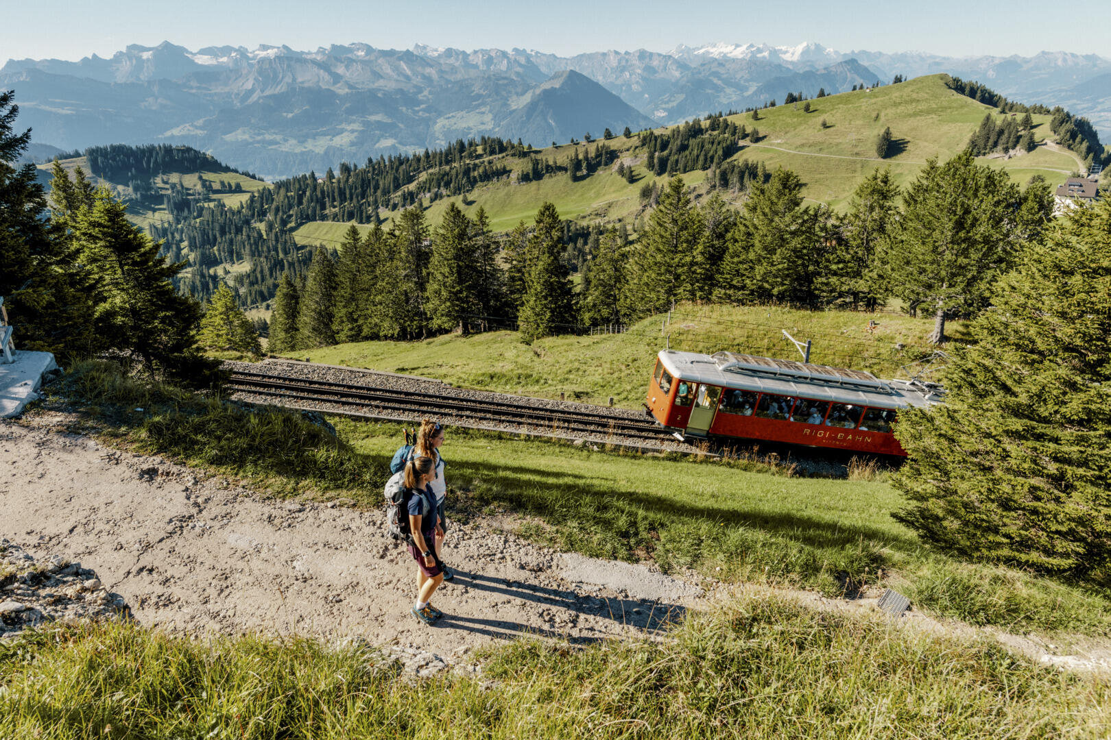Ein Wanderer steht auf einem Bergpfad mit einer roten Zahnradbahn, die durch üppig grüne Hügel und Pinienwälder führt, mit fernen schneebedeckten Bergen unter einem klaren blauen Himmel.