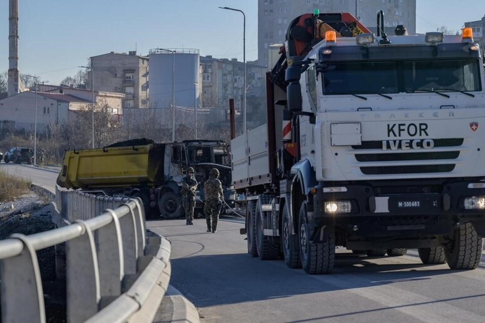Ein IVECO-Lkw der KFOR steht auf einer Straße neben einem ausgebrannten gelben Kipplaster. Zwei Uniformierte stehen zwischen den Fahrzeugen. Im Hintergrund sind Gebäude und Industrieanlagen zu sehen.