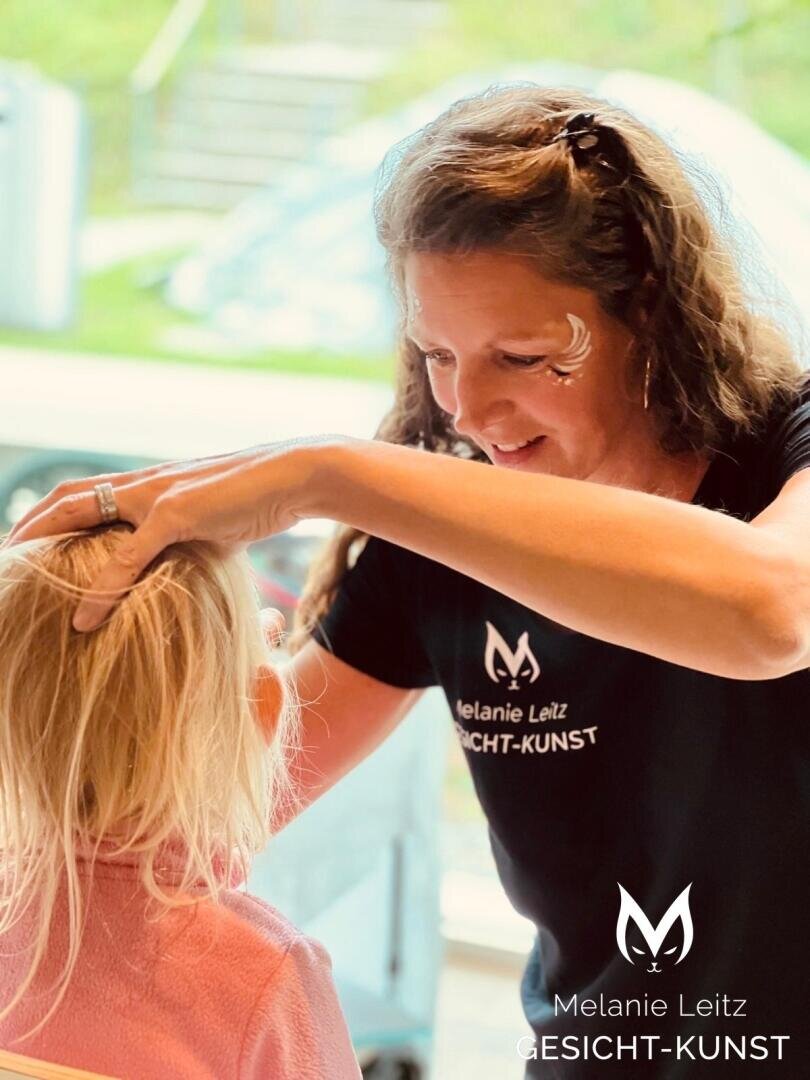 Eine Frau mit Gesichtsbemalung und einem Melanie Leitz GESICHT-KUNST-Shirt bemalt sanft das Gesicht eines Kindes mit blondem Haar, beide in einer hellen Innenumgebung.