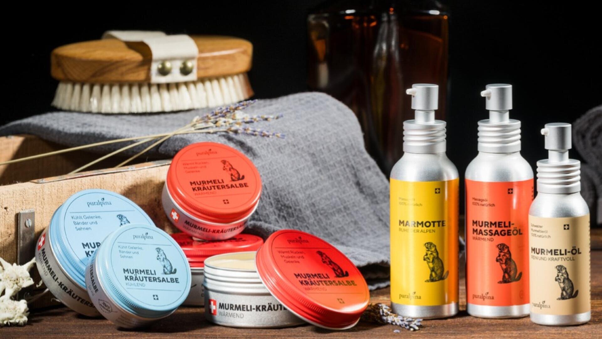 Eine Auswahl von Murmeltierölprodukten und Kräuterbalsamen in Dosen und Flaschen ist auf einem Tisch mit einer Holzbürste, einem grauen Handtuch, getrockneten Blumen und Glasflaschen im Hintergrund angeordnet.