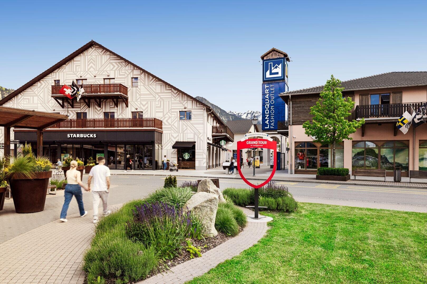 Ein Mann und eine Frau gehen auf ein Gebäude mit einem Starbucks zu. In der Nähe befindet sich ein blaues Schild für das Landquart Fashion Outlet und eine grüne Landschaft unter einem klaren blauen Himmel.