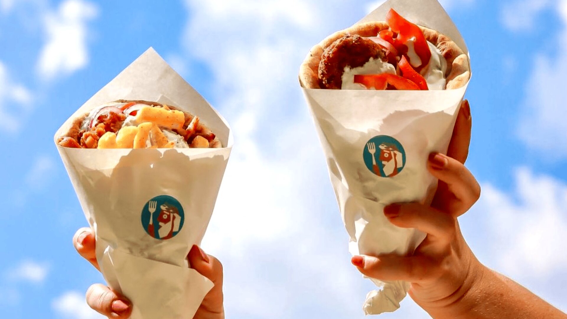 Zwei Hände halten mit Falafel, Gemüse und Soße gefüllte Pita-Wraps, eingewickelt in weißes Papier mit einem Restaurantlogo, vor einem strahlend blauen Himmel mit vereinzelten Wolken.