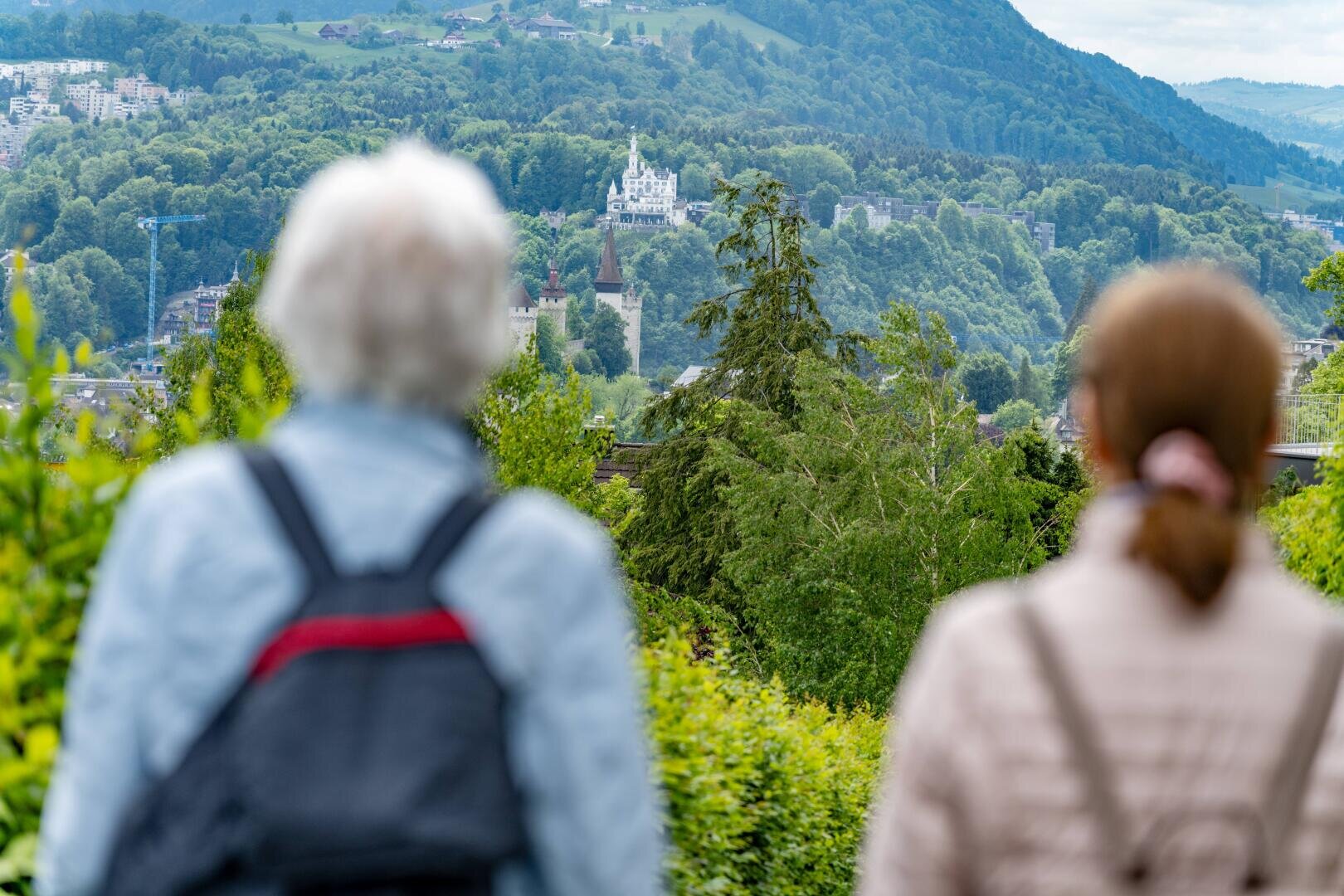 Zwei Personen mit Rucksäcken blicken über eine üppige grüne Landschaft auf ein entferntes weißes Schloss auf einem Hügel, umgeben von Bäumen und Bergen, unter einem teilweise bewölkten Himmel.