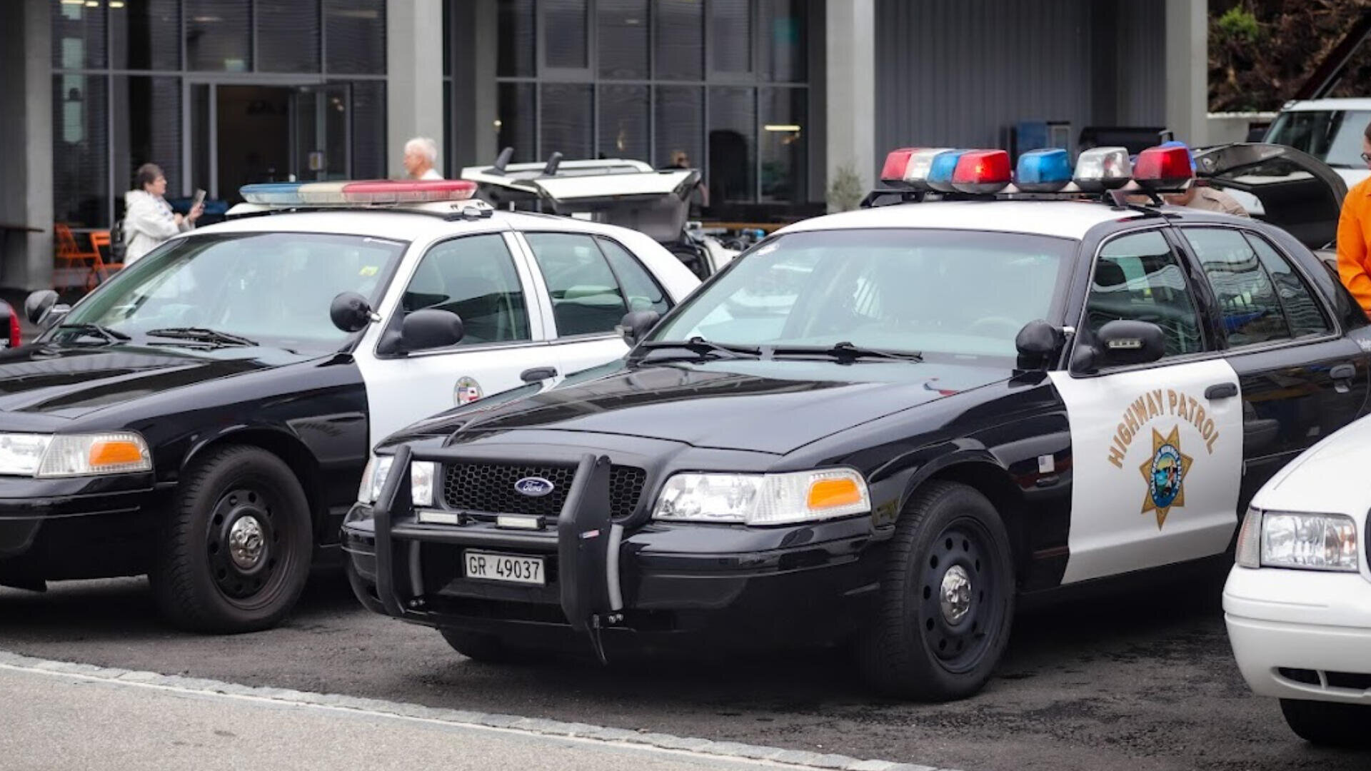 Zwei schwarz-weiße Polizeiautos mit Dachlampen sind nebeneinander vor einem Gebäude geparkt. Ein Wagen ist mit "Highway Patrol" gekennzeichnet. Im Hintergrund sind Menschen zu sehen.