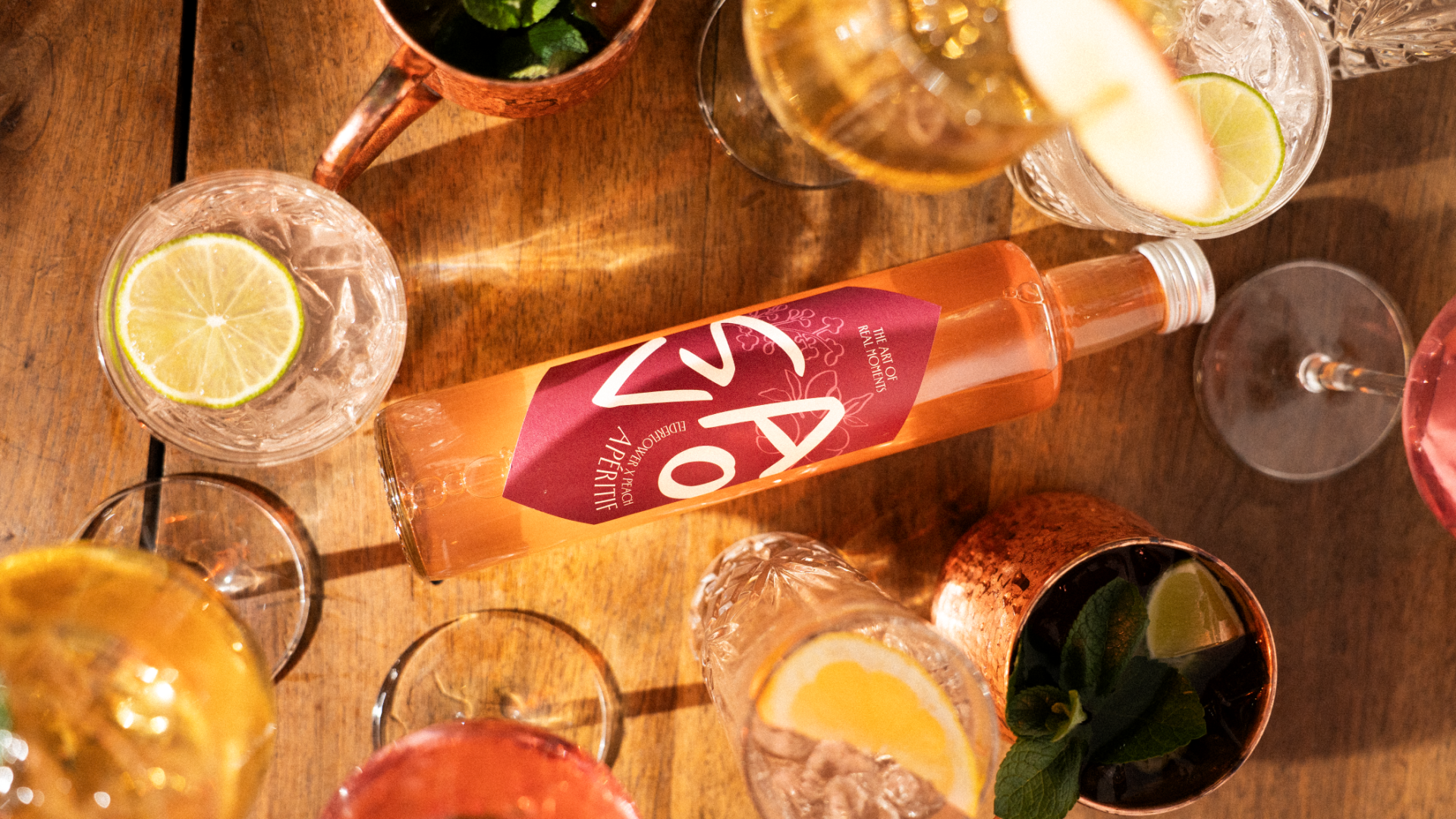 Eine Flasche Ao Aperitif steht auf einem Holztisch, umgeben von verschiedenen Cocktails in Glas- und Kupferbechern, garniert mit Limette, Zitrone und Minze. Das Sonnenlicht schafft eine warme, einladende Atmosphäre.