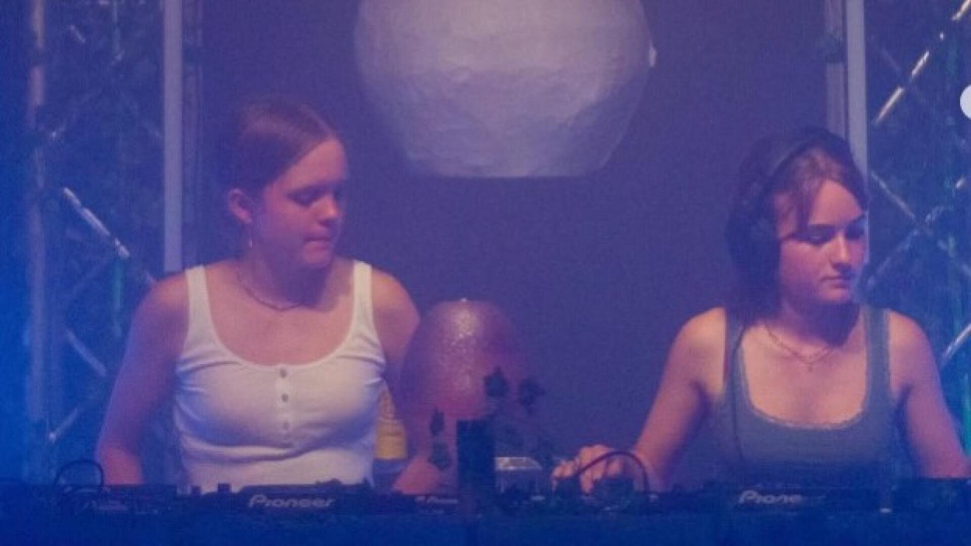 Zwei junge Frauen legen gemeinsam auf der Bühne auf und stehen nebeneinander hinter dem DJ-Equipment. Beide sind konzentriert, wobei eine Kopfhörer trägt. Der Hintergrund ist schwach beleuchtet, mit lila-blauer Beleuchtung und sichtbaren Bühnenstrukturen.