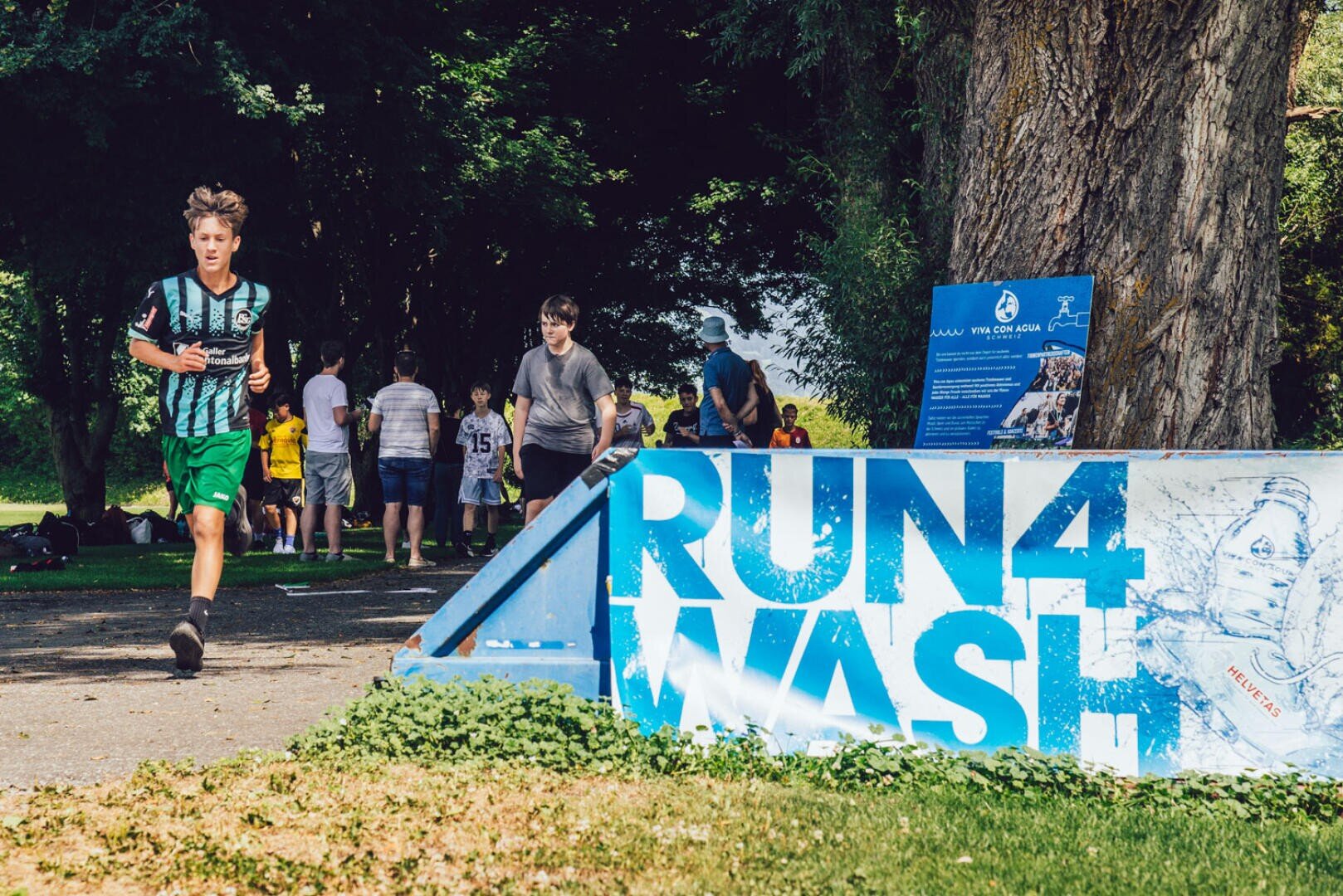 Ein Junge in Sportkleidung rennt an einem großen Schild mit der Aufschrift "RUN 4 WASH" in der Nähe eines Baumes vorbei, während sich im Hintergrund andere Kinder und Erwachsene an einem sonnigen Tag versammeln.