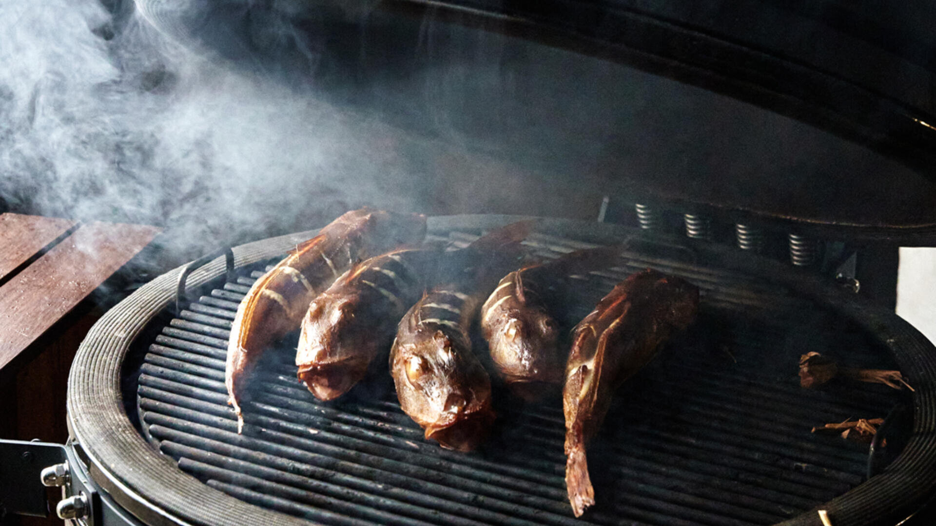 Vier ganze Fische werden auf einem Grill gegrillt, um die herum Rauch aufsteigt. Der Grilldeckel ist geöffnet, und die Fische sehen verkohlt und gar aus. Links ist teilweise ein Holztisch zu sehen.