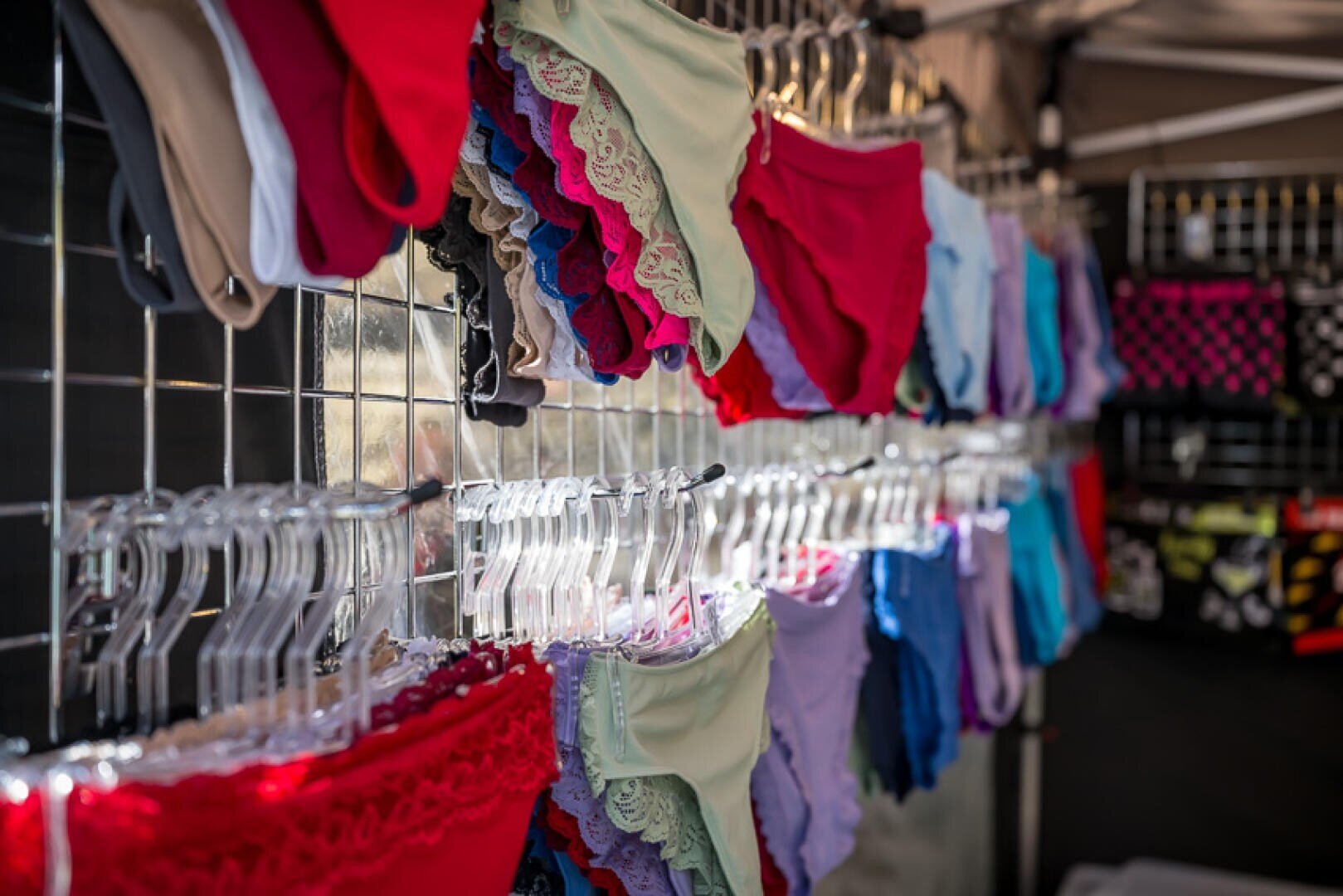 An einem Marktstand wird bunte Damenunterwäsche, darunter Slips und Höschen mit Spitzenbesatz, auf durchsichtigen Plastikbügeln und Metallgestellen ausgestellt. Im Hintergrund sind weitere Unterwäsche und Accessoires zu sehen.