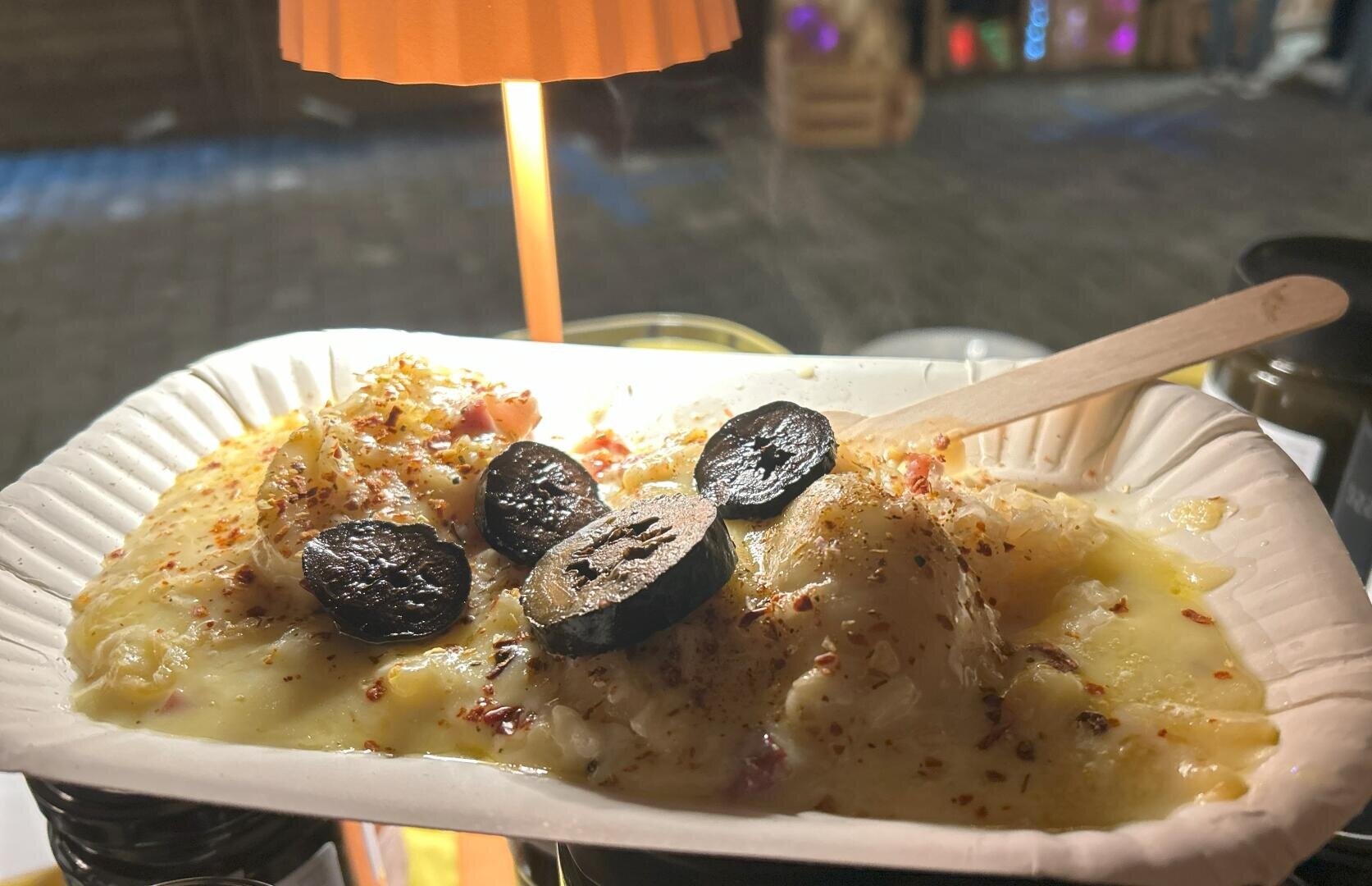 Auf einem Pappteller liegt geschmolzener Käse über Kartoffeln, die mit Wurstscheiben belegt und mit Gewürzen bestreut sind. Eine Holzgabel liegt auf dem Essen, im Hintergrund scheint das warme Licht einer Lampe.