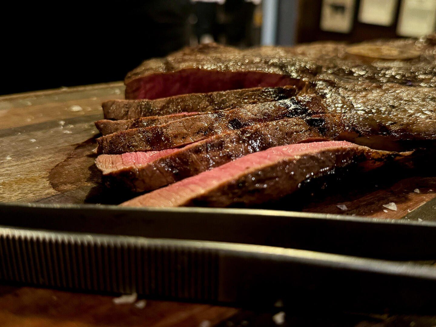Nahaufnahme eines in Scheiben geschnittenen, gegrillten Steaks auf einem hölzernen Schneidebrett, das ein saftiges, rosafarbenes Inneres und eine angebratene, braune Außenseite zeigt. Im Vordergrund ruht ein Metallmesser.