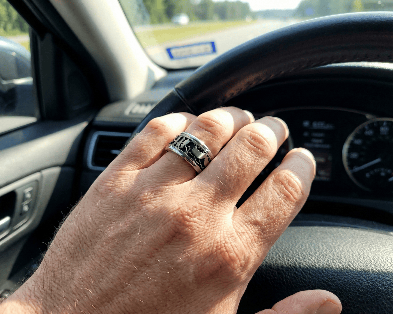 Eine Nahaufnahme einer Hand mit einem silbernen Zierring, die auf einem Autolenkrad ruht. Das Sonnenlicht beleuchtet die Hand, und durch die Windschutzscheibe im Hintergrund sind eine Straße und Bäume zu sehen.