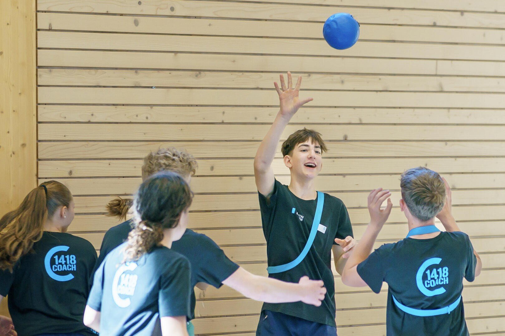 Eine Gruppe Jugendlicher in schwarzen Hemden spielt ein Hallenballspiel. Ein Junge streckt einen blauen Ball in die Höhe, um ihn zu fangen oder zu werfen, während andere, auf deren Hemden "1418 COACH" steht, zusehen oder mitmachen. Die Kulisse besteht aus Holzwänden.