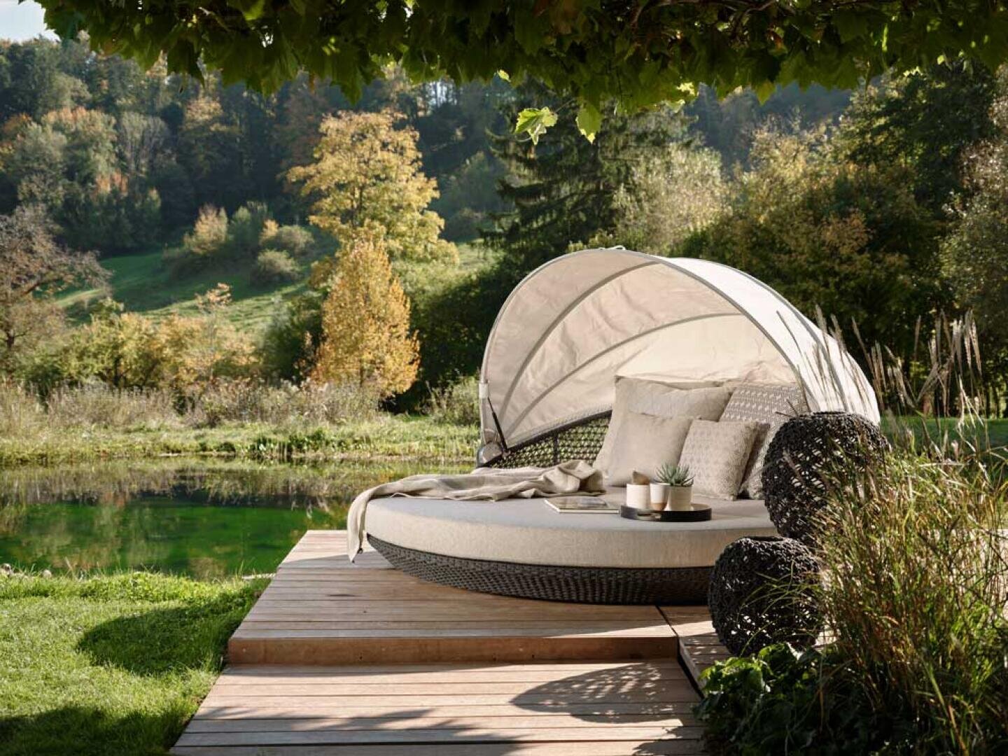 Eine runde Liege mit Baldachin und Plüschkissen steht auf einer Holzterrasse an einem Teich, umgeben von üppigem Grün und Bäumen unter einem belaubten Ast. Auf einem kleinen Tisch stehen eine Pflanze und eine Tasse.