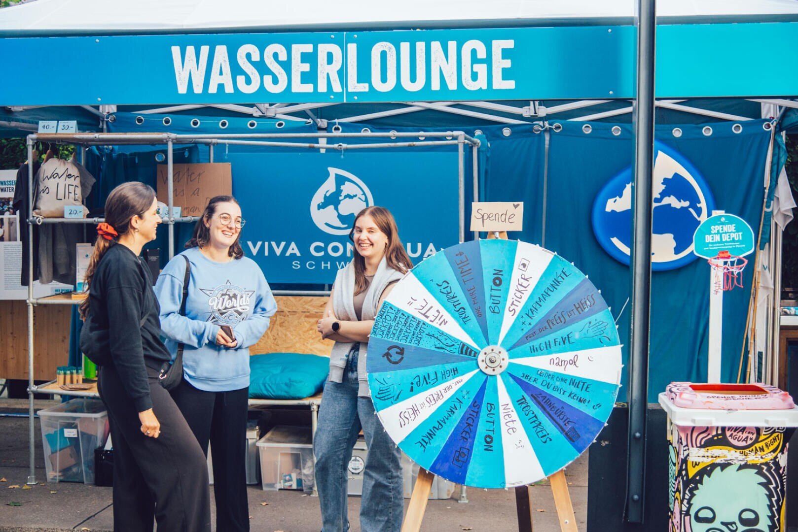 Drei Frauen stehen vor einem blauen Wasser Lounge-Stand, lächeln und unterhalten sich in der Nähe eines bunten, sich drehenden Preisrades, während im Hintergrund Spenden- und Informationsschilder zu sehen sind.