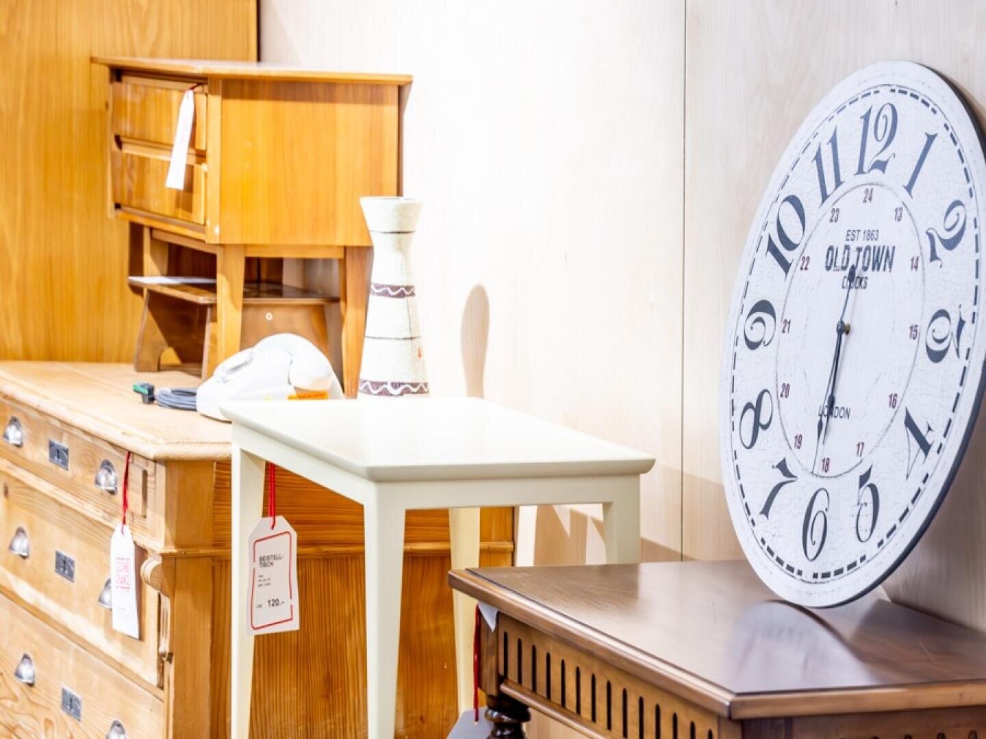 Ein weißer Beistelltisch mit einer Vase und einer Mütze darauf steht zwischen zwei hölzernen Kommoden. Eine große Uhr im Vintage-Stil lehnt an der Wand. An Tisch und Kommode hängen Preisschilder.