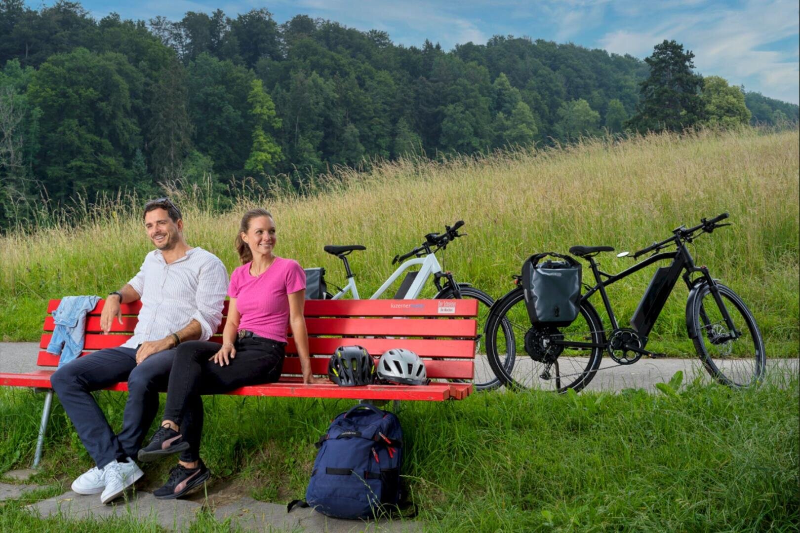 Ein Mann und eine Frau sitzen auf einer roten Bank auf einer Wiese, hinter ihnen sind zwei Fahrräder abgestellt. Helme, ein Rucksack und eine Jacke liegen auf der Bank und im Gras. Im Hintergrund sind Bäume und ein blauer Himmel zu sehen.