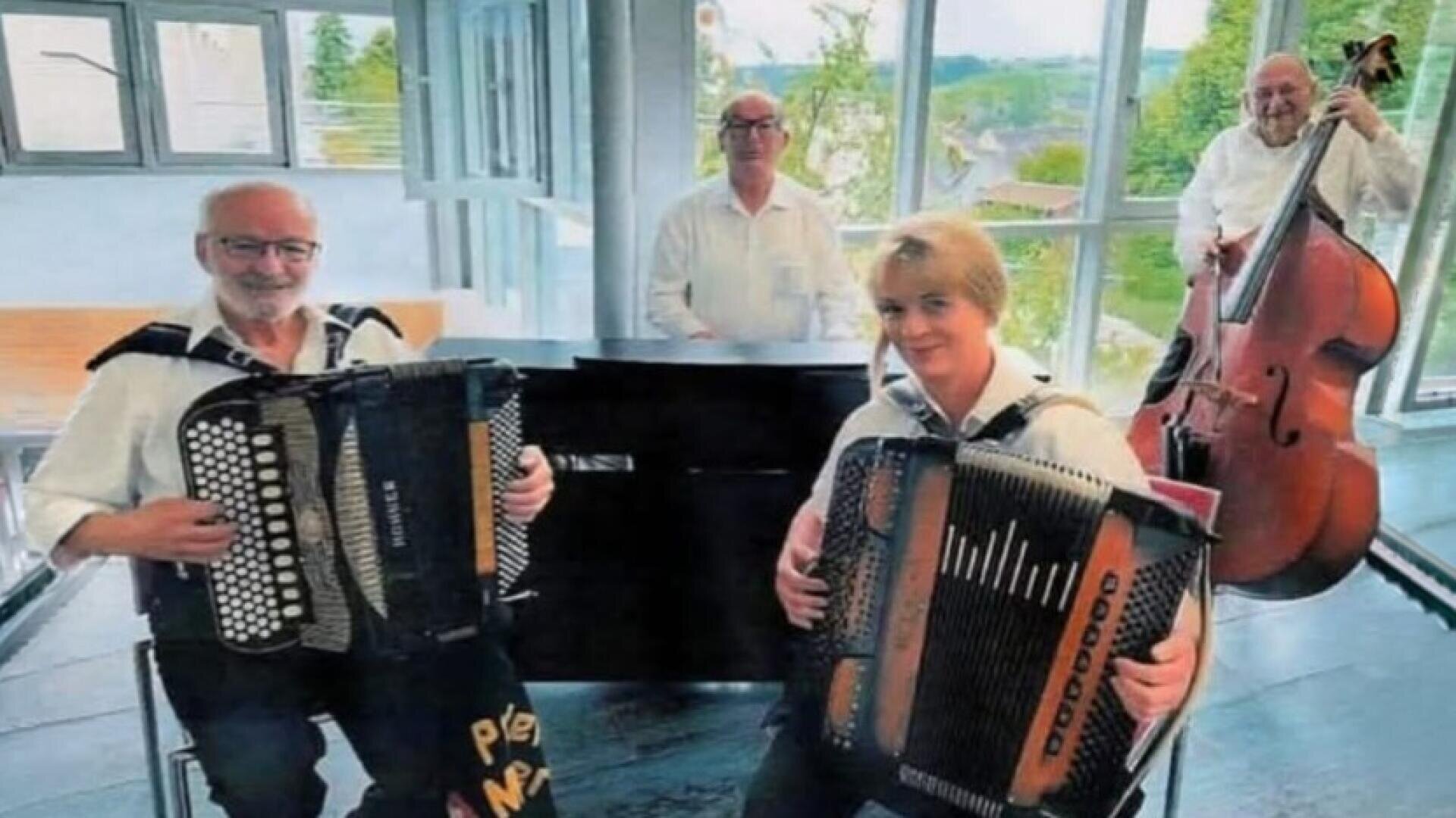 Vier Musiker in weißen Hemden treten in einem Innenraum mit großen Fenstern hinter ihnen auf. Zwei von ihnen spielen vorne Akkordeon, während die anderen beiden hinten Klavier und Kontrabass spielen. Die Szene ist hell und fröhlich.
