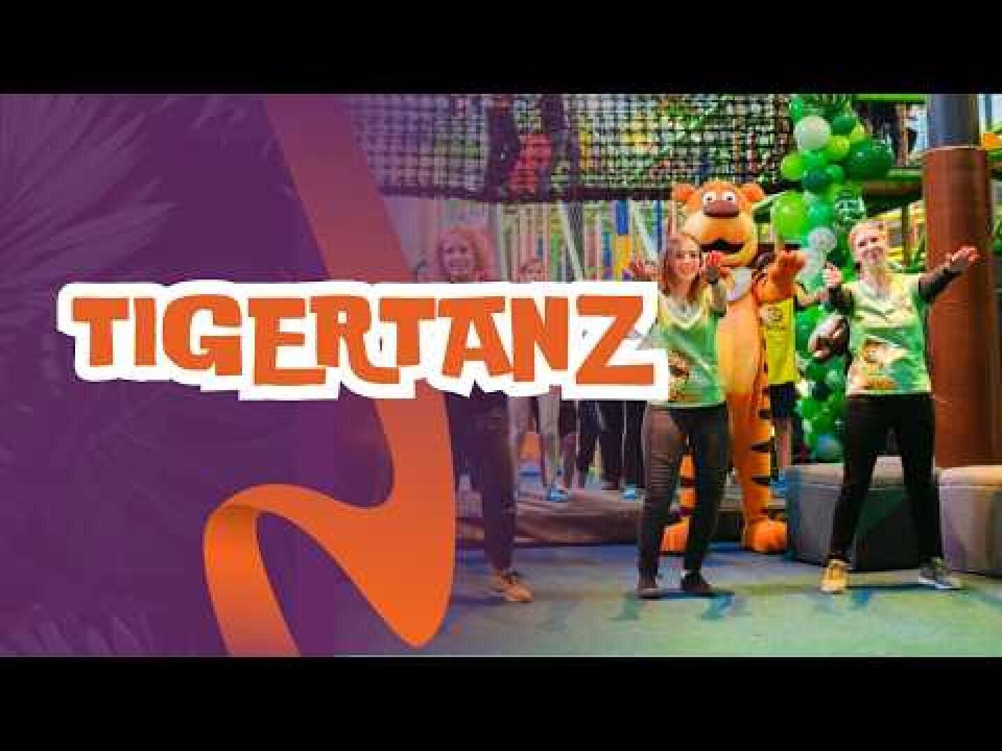 Vier Personen und eine Person in einem Tigerkostüm tanzen in einer farbenfrohen Innenumgebung mit grünen Luftballons, unter dem fetten Text TIGERTANZ auf einem lila und orangefarbenen Hintergrund.