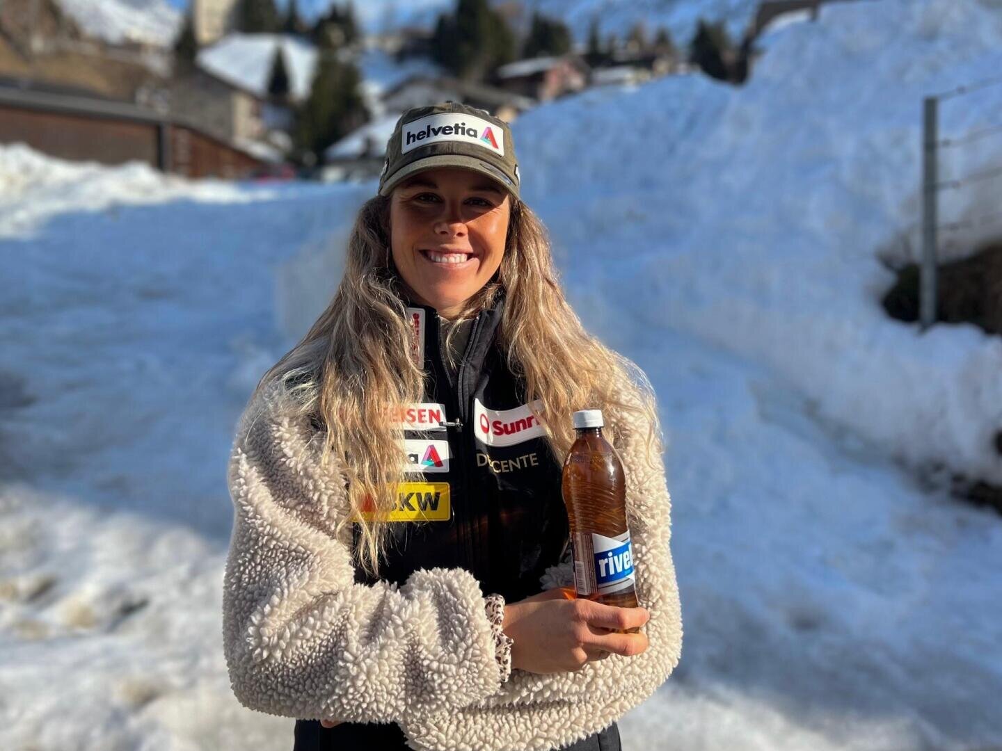 Eine lächelnde Frau in Winterjacke und Markenmütze steht draußen in einer verschneiten Landschaft und hält eine Flasche mit der Aufschrift riva. Die Sonne scheint und wirft Schatten auf den Schnee hinter ihr.