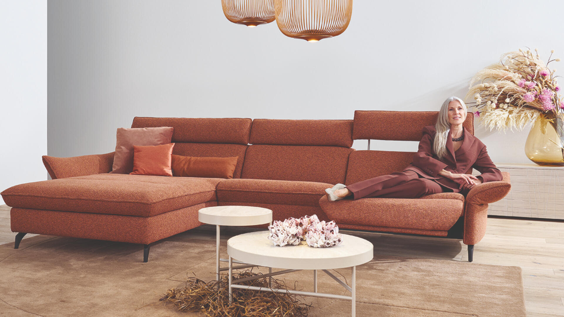 Eine Frau in einem braunen Anzug sitzt auf einem großen rostfarbenen Sektionssofa mit mehreren passenden Kissen, neben runden Couchtischen und einer Vase mit Trockenblumen in einem modernen, minimalistischen Wohnzimmer.