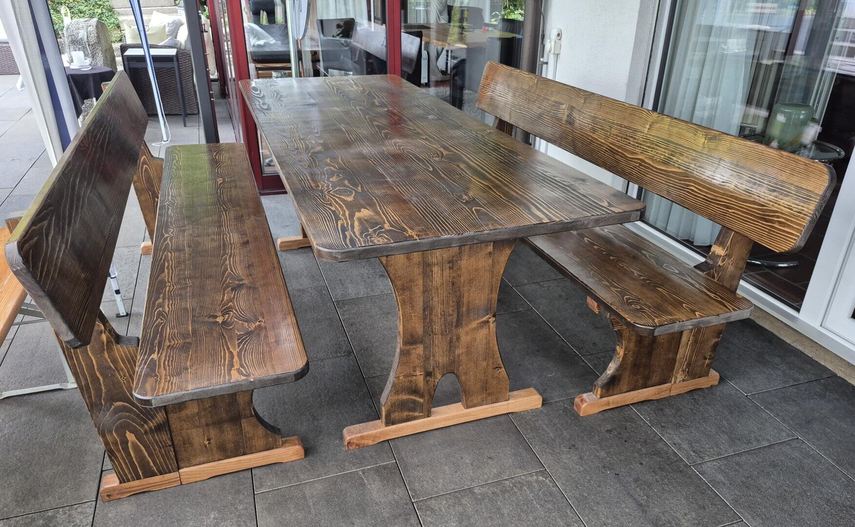 Ein hölzerner Esstisch für den Außenbereich mit zwei passenden Bänken auf beiden Seiten, auf einer gefliesten Terrasse in der Nähe von Glastüren und umgeben von Gartenmöbeln.