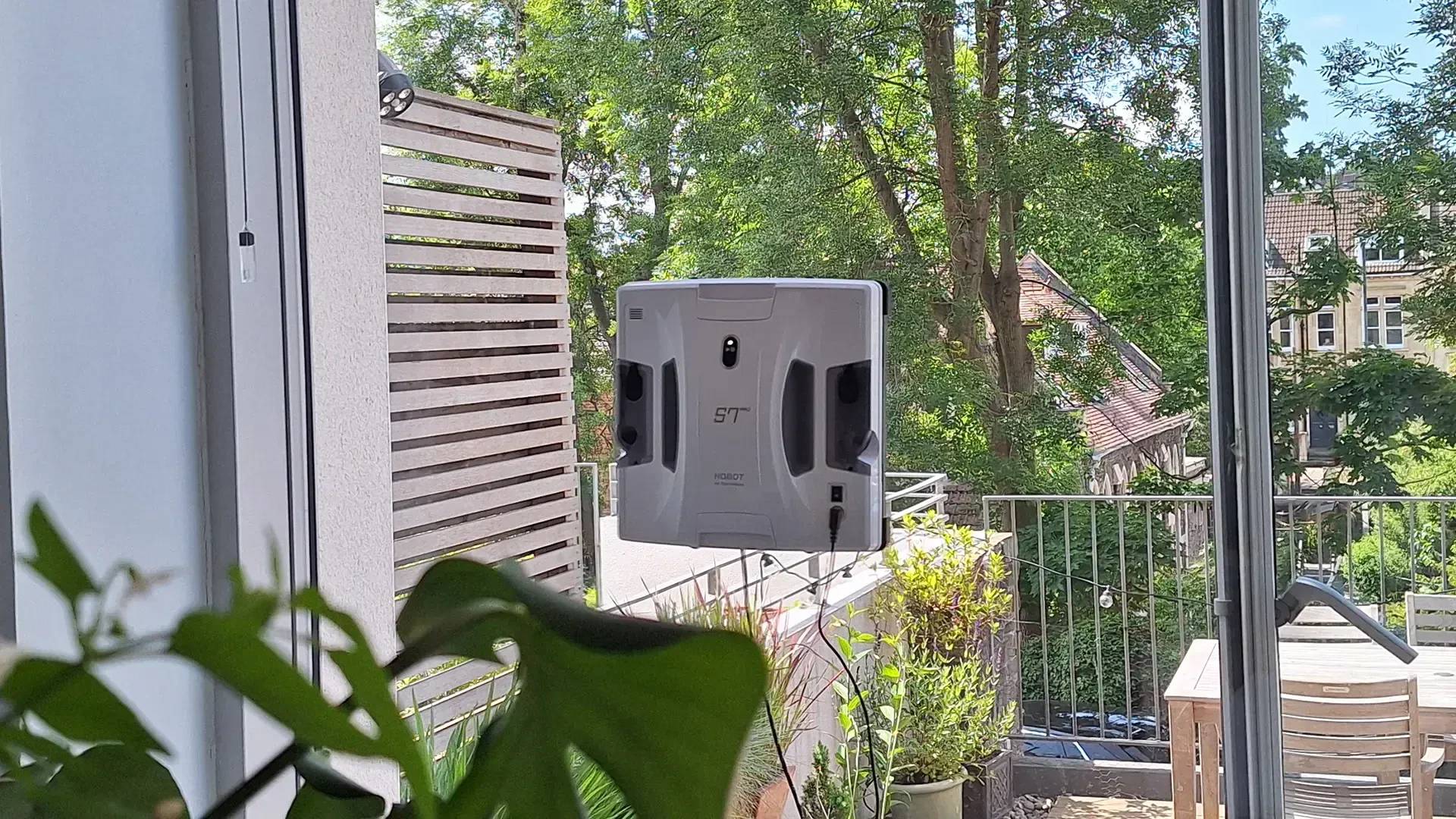 Ein Fensterreinigungsroboter ist an einem großen Fenster angebracht und reinigt dessen Oberfläche. Im Hintergrund sind Grünpflanzen und eine Außenterrasse mit Tisch und Stühlen an einem sonnigen Tag zu sehen.